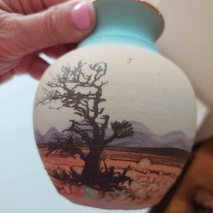 May include: A small ceramic vase with a rounded body and a light blue neck featuring a gold rim. The surface displays a desert landscape illustration with a dark, leafless tree in the foreground, distant purple mountains, and a warm orange horizon.