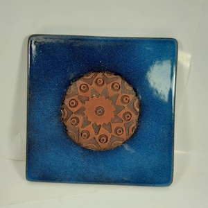 Puede incluir: Azulejo cerámico cuadrado con un esmalte azul profundo y brillante. El centro presenta un diseño circular en relieve en tonos marrones, con un patrón repetitivo de círculos y formas geométricas. El azulejo mide aproximadamente 15 cm cuadrados.
