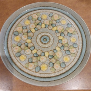 May include: Round ceramic plate with a floral design. The plate features a light blue rim and a central pattern of yellow, green, and white flowers arranged in a circular pattern. The plate has a textured surface and a neutral color palette.