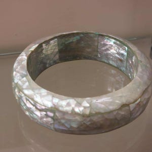 May include: A wide bangle bracelet constructed from numerous small, iridescent mother-of-pearl shell tiles. The tiles are arranged in a mosaic pattern, displaying a spectrum of white, silver, and soft pastel colors with a reflective, glossy finish.