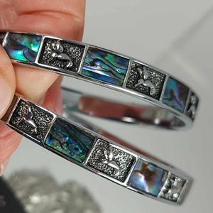 May include: Two silver bangles featuring rectangular abalone shell inlays and hummingbird motifs. The iridescent shell segments display blue, green, and purple hues. The bangles are held by a hand, reflecting light.