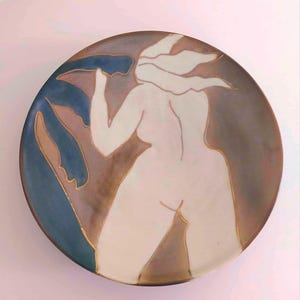 Puede incluir: Un plato de cerámica redondo que presenta una figura blanca estilizada de una persona con cabello suelto, delineada en oro. El fondo consiste en formas de hojas azul oscuro y un degradado marrón, creando contraste con la figura central.