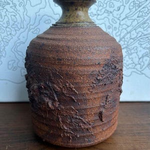 May include: A rustic brown ceramic vase with a textured surface and a wide, rounded base. The vase has a slightly flared opening and a brown glaze.
