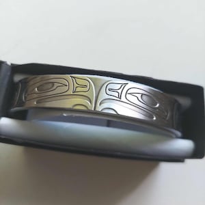 Peut inclure: Un bracelet manchette en métal argenté orné d'une gravure de style formline autochtone de la côte Nord-Ouest représentant un œil et des formes abstraites. Le bracelet est présenté dans une boîte rectangulaire noire avec un insert intérieur blanc.