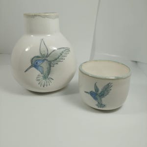 May include: A white ceramic vase and cup set, each adorned with a hand-painted hummingbird design in blue and green. The vase has a rounded shape and a narrow neck. The cup is cylindrical with a matching hummingbird motif.