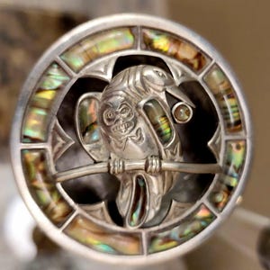 May include: A circular silver brooch featuring a central carved raven figure perched on a branch, accented with iridescent abalone shell inlays. The outer frame is segmented with rectangular pieces of abalone, creating a decorative border.