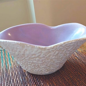 May include: A heart-shaped ceramic bowl featuring a smooth, light lavender interior and a textured, matte white exterior. The bowl has a distinct, irregular rim that follows the heart shape, creating a curved and organic silhouette for the vessel.