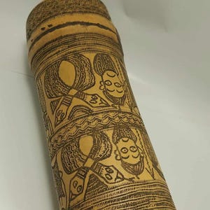 Puede incluir: Un cilindro de bambú marrón con intrincados diseños grabados en negro de rostros estilizados y patrones. El cilindro mide aproximadamente 15 cm de alto.