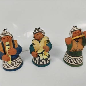 Può includere: Tre piccole figurine in ceramica di persone che suonano strumenti musicali. Le figurine indossano abiti tradizionali e hanno la pelle marrone. La figura a sinistra suona il flauto, la figura al centro suona il tamburo e la figura a destra suona un flauto lungo.
