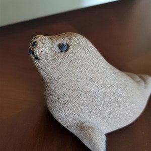 Op de afbeelding: Een klein, zandkleurig keramiek zeehondbeeldje met zwarte ogen. De zeehond kijkt omhoog en heeft een licht geopende mond.