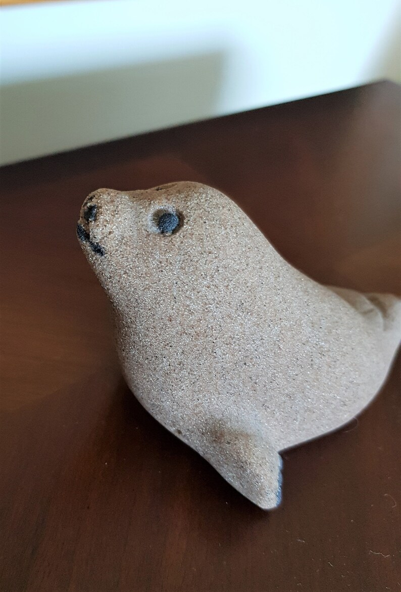 Op de afbeelding: Een klein, zandkleurig keramiek zeehondbeeldje met zwarte ogen. De zeehond kijkt omhoog en heeft een licht geopende mond.