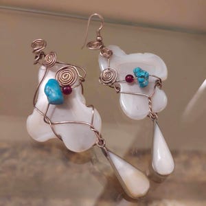 Op de afbeelding: Een paar witte schelp oorbellen met koperdraad eromheen gewikkeld. De oorbellen zijn versierd met turquoise en rode kralen. De oorbellen hebben een traanvorm.