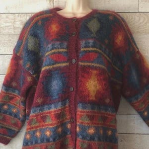 Könnte beinhalten: Knopf-Cardigan mit farbenfrohem geometrischem Muster. Der Pullover hat eine Mischung aus roten, blauen, gelben und grünen Farbtönen. Das Design umfasst Rauten und Streifen. Der Pullover hat einen runden Ausschnitt und lange Ärmel.