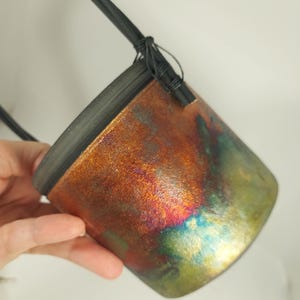 Könnte beinhalten: Kleiner, metallisch aussehender Behälter mit schwarzem Rand und Riemen. Der Behälter hat eine strukturierte Oberfläche mit einer Mischung aus Orange, Rot, Grün und Blau. Der Riemen ist schwarz und mit Draht am Behälter befestigt.