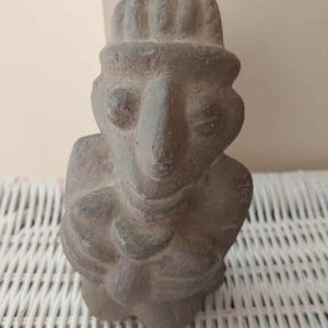 May include: A gray, textured clay figurine depicting a stylized figure with a prominent nose, almond-shaped eyes, and a rounded hat. The figure has its arms crossed over its chest, with a smooth, matte finish and visible surface imperfections.