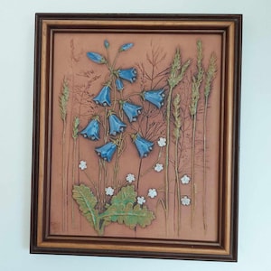 May include: A framed ceramic wall art piece depicting bluebell flowers and wheat stalks in a brown and green colour scheme.