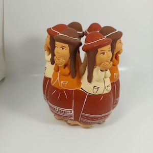 Op de afbeelding: Een keramieken beeldje met zes figuren in een cirkel. Elke figuur draagt een hoed en een lange jurk. De jurken zijn roodbruin met witte accenten. De figuren hebben een gebruinde huid, bruin haar en dragen oranje en crèmekleurige tops.