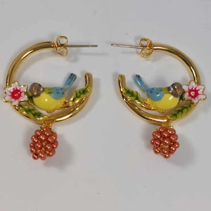 Peut inclure: Boucles d'oreilles créoles dorées avec un oiseau bleu perché sur une branche verte avec une fleur rose. Les boucles d'oreilles ont un pendentif en forme de grappe de perles orange.