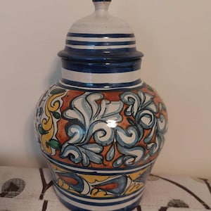 May include: A ceramic jar with a lid, featuring a vibrant design. The jar has a white and blue striped lid, a blue rim, and a body adorned with orange, yellow, and blue floral patterns. The jar is a decorative piece.