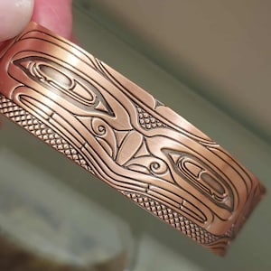 Peut inclure: Bracelet manchette de couleur cuivre avec un motif en relief complexe. Le bracelet présente un motif répétitif de formes et de lignes stylisées, avec une bordure texturée. Le métal a une teinte brun-rouge chaud.