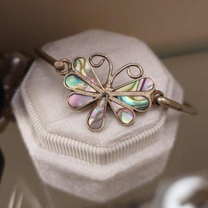 Peut inclure: Un bracelet en métal argenté avec un ornement central en forme de papillon. Les ailes du papillon sont incrustées de segments de nacre iridescente affichant des nuances de vert, de bleu et de violet. La monture métallique est polie.