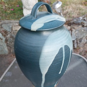 May include: A handcrafted ceramic jar with a lid and handle. The jar is a deep teal color with white dripped accents. The lid has a matching teal and white design. The jar is approximately 12 inches tall.