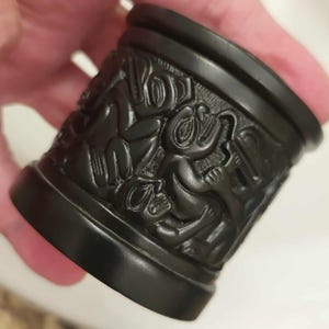May include: A cylindrical black object featuring intricate, carved relief patterns of stylised animal figures wrapping around the exterior surface. The piece has a smooth, matte finish with a raised rim at the top and a solid base at the bottom.
