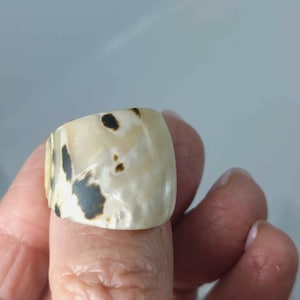 May include: A wide, smooth ring featuring a cream-coloured base with irregular, dark brown to black mottled spots and patches distributed across its surface. The band has a polished finish and a rounded, chunky silhouette that encircles the finger.