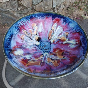 May include: A round ceramic bowl featuring a vibrant glaze with deep blue, magenta, and purple hues. The interior surface displays a swirling, marbled pattern with streaks of white and tan radiating from the centre toward the dark blue rim.