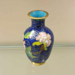 May include: A small cloisonné vase with a deep blue background featuring pink and white floral motifs and green leaves. The vase has a gold-toned metal rim and base, with a bright turquoise interior and delicate gold wire outlines throughout the design.