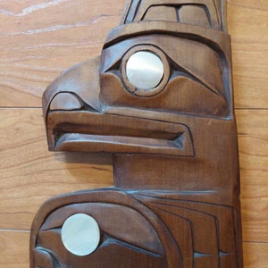 Puede incluir: Una talla de madera de un tótem nativo americano con cabeza de pájaro y dos ojos hechos de concha blanca. El tótem es marrón y tiene tallas intrincadas.