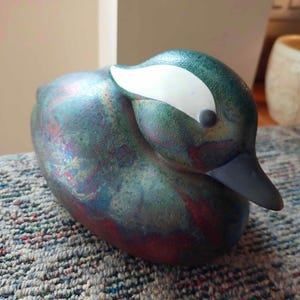 May include: A duck figurine featuring a textured surface with a mottled finish in shades of green, blue, and red. The duck has a smooth white patch on the side of its head, a dark grey beak, and a small black eye, presented in a resting position.