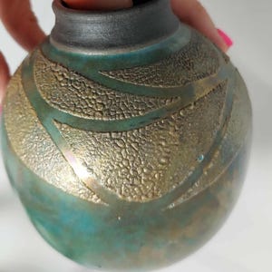 May include: A small, round ceramic vase with a matte black rim and a spherical body. The surface features a textured, metallic gold leaf pattern over a deep teal and bronze glaze, creating a contrast between smooth and rough finishes.