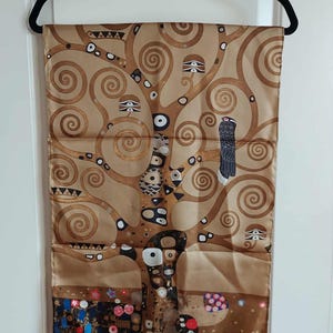 May include: A rectangular silk scarf featuring a gold and brown tree of life motif with swirling branches, geometric patterns, and a dark bird perched on a limb. The lower section displays colourful floral accents against a dark brown base.