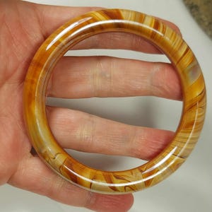 May include: A circular amber-coloured bangle bracelet with a marbled pattern of light and dark brown hues. The bracelet is smooth and polished, reflecting light. The jewellery piece is held in a hand.
