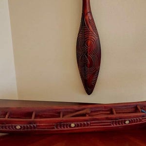 Könnte beinhalten: Ein geschnitztes Holzkajak in einem tiefen Rotbraun mit aufwendigen Designs und einem fischförmigen Detail am Heck. Ein passender Paddel hängt über dem Kajak. Das Kajak wird auf einer Holzoberfläche präsentiert. Das Design deutet auf kulturelle Bedeutung hin.