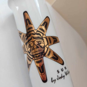 Peut inclure: Vase en céramique blanche orné d'un motif de soleil stylisé noir et doré. Le soleil a un visage et des rayons. Le texte "SUN by Jody Wilson" est imprimé en noir sur le vase.