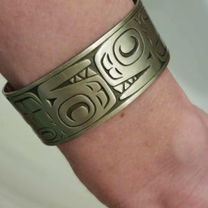 May include: A silver-toned cuff bracelet with intricate, engraved Native American-inspired designs. The bracelet features stylized animal and geometric patterns. The metal has a slightly aged appearance, enhancing the detail of the artwork. The cuff is wide and wraps around the wrist.