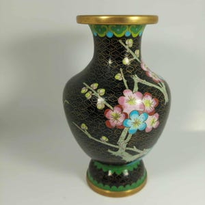 May include: A black cloisonné vase with a gold rim and base. The vase features a floral design with pink and blue blossoms on a branch. The vase has a bulbous shape and a narrow neck, standing approximately 15 cm tall.
