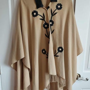 May include: A tan poncho featuring a black leather collar and black floral applique detailing on the front. The garment has a single button closure at the neckline and a fringe hemline along the bottom edge, providing a structured yet draped appearance.