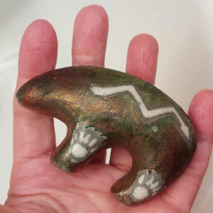 May include: A bear-shaped stone carving with a metallic bronze and green patina finish. The figure features inlaid white stone details, including a zigzag line across the back and paw prints on the feet, creating a distinct contrast in texture.