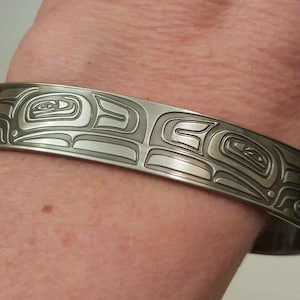 May include: A silver-coloured cuff bracelet with an engraved design. The bracelet features a repeating pattern of stylized shapes and lines, possibly representing a cultural or artistic motif. The metal has a matte finish, and the bracelet is worn on a wrist.