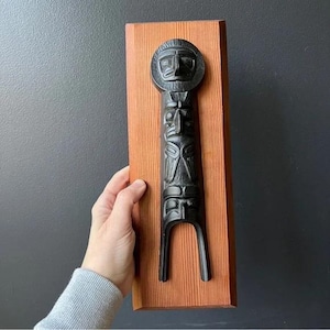 May include: A black carved wooden totem pole mounted on a rectangular, reddish-brown wooden board. The totem pole features several carved faces and figures. The piece is held by a hand against a dark gray background.