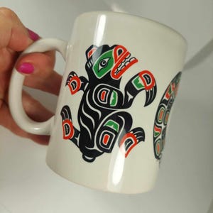 Puede incluir: Taza de cerámica blanca con un diseño estilizado en negro, rojo y verde de un oso. El oso está de pie con la boca abierta y las garras extendidas.