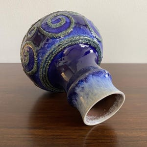 May include: A spherical ceramic vase featuring a deep cobalt blue glaze with textured, raised green spiral patterns across the body. The neck transitions into a lighter, creamy off-white glaze at the rim, which has a smooth, flared opening.