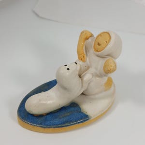 May include: A ceramic figurine of a person in a hooded parka hugging a seal. The figure is mainly off-white with tan details, set on a blue and yellow base. The seal has black dot eyes.