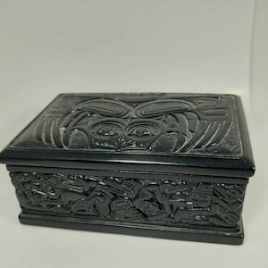 May include: Black wooden box with intricate carvings of a stylized animal face and other designs. The box is closed.
