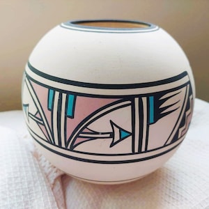 Pode incluir: Um vaso redondo de cerâmica esbranquiçada com uma borda preta e uma faixa decorativa com desenhos geométricos em rosa, turquesa e preto. O vaso tem uma superfície lisa e uma pequena abertura no topo. O design lembra a cerâmica nativa americana.