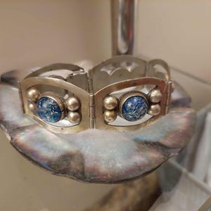 Peut inclure: Un bracelet argenté à charnière. Le bracelet présente des pierres bleues ovales avec un motif moucheté, encadrées par une monture argentée. De petits accents argentés ronds sont placés autour des pierres. Le bracelet est présenté sur une surface nacrée.
