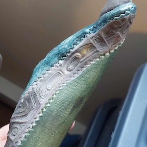 May include: A green and grey carved wooden sculpture of a stylised animal with a textured surface. The sculpture has a raised design of a stylised animal with a geometric pattern.